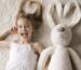 Child in a white dress lying on a cream rug next to a large white plush rabbit.