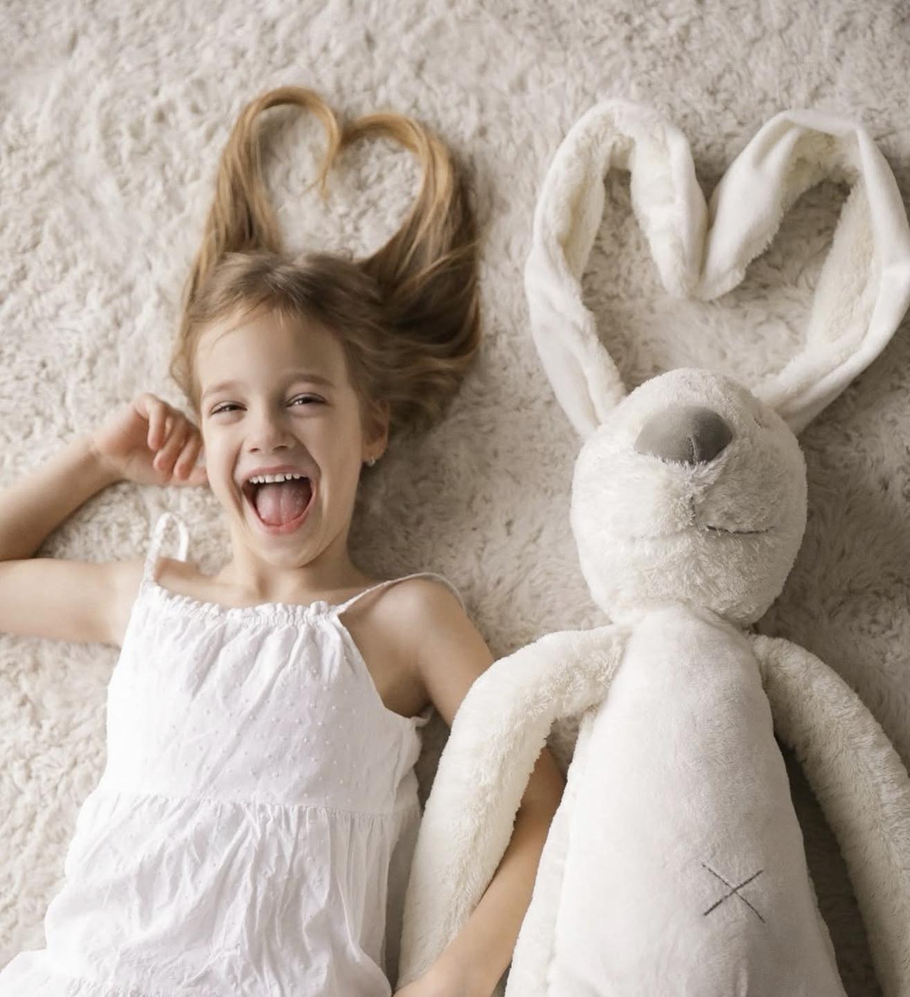 Child in a white dress lying on a cream rug next to a large white plush rabbit.