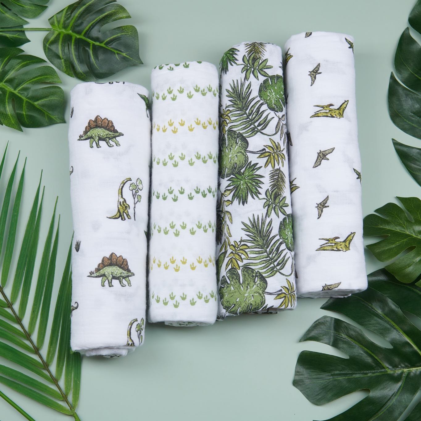Four rolled white baby cloths with green dinosaur and leaf prints arranged among large green leaves.