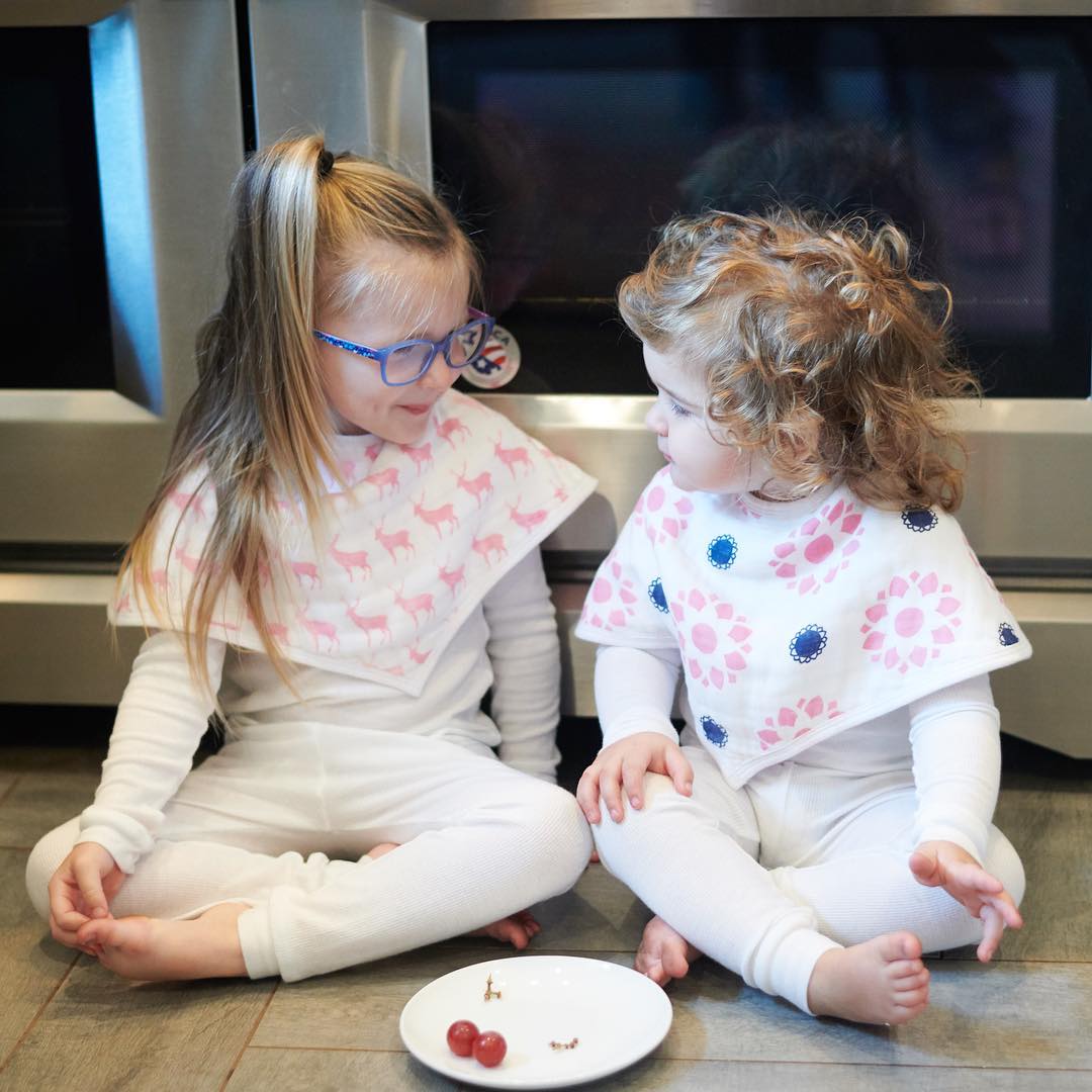 Two children in white outfits sit on a kitchen floor wearing patterned bibs beside a white plate with two grapes.